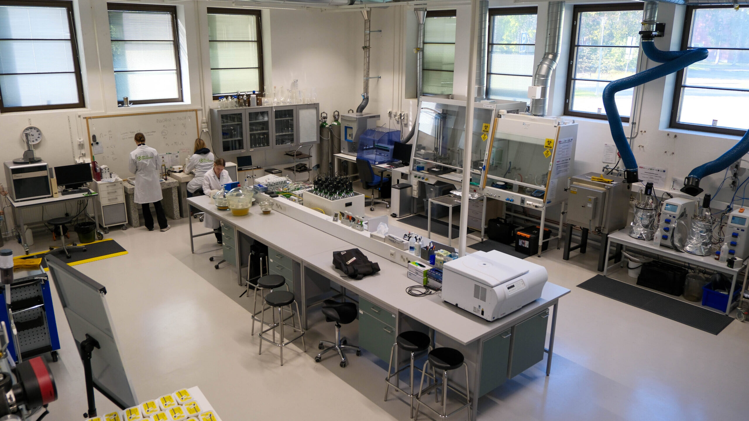 Moderni laboratorio, jossa kaksi valkoisiin laboratoriotakkeihin pukeutunutta henkilöä työskentelee tiskin ääressä. Huoneessa on laboratoriolaitteita, tietokoneita, jakkaroita, suuret ikkunat ja siniset poistoilmaventtiilit, ja penkkien päällä on erilaisia tieteellisiä välineitä ja tarvikkeita.